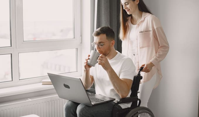 A support worker sharing coffee with a man in a wheelchair while he works on a laptop